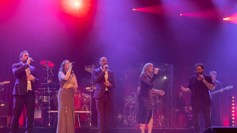 Fünf Sänger performen auf einer Bühne, umgeben von bunten Lichtern. Eine Band spielt im Hintergrund. Die Atmosphäre ist festlich und lebendig., © Schönblick gemeinnützige GmbH