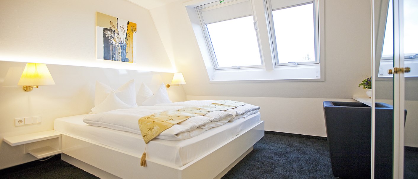 Helles Schlafzimmer mit Doppelbett, zwei Dachfenstern, Wandkunst und Nachttischlampen. Der Raum wirkt modern und einladend., © Hotel Krone Helles Schlafzimmer mit Doppelbett, zwei Dachfenstern, Wandkunst und Nachttischlampen. Der Raum wirkt modern und einladend., © Hotel Krone