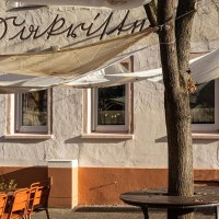 Ein Caf&eacute; mit Sonnensegeln und orangenen St&uuml;hlen im Au&szlig;enbereich. Die Fassade ist beige mit Schriftzug. Ein Baum steht vor dem Eingang., &copy; Stuttgart-Marketing GmbH, Sarah Schmid