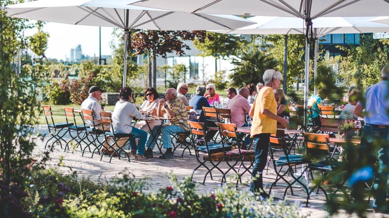 Menschen sitzen unter gro&szlig;en Sonnenschirmen an Holztischen im Freien, umgeben von gr&uuml;ner Vegetation und Blumen., &copy; Wilhelm Kern GmbH