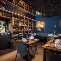 Stilvolles Restaurant mit gedeckten Tischen, Weinregalen und moderner Beleuchtung. Eine offene K&uuml;che und ein Kunstwerk an der Wand sind sichtbar., &copy; Belser