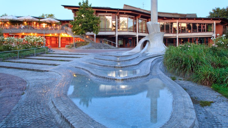 Moderner Brunnen mit geschwungenen Formen und Wasserbecken vor einem beleuchteten Geb&auml;ude mit Terrasse und Pflanzen., &copy; Waiblingen - Stuttgart-Marketing GmbH