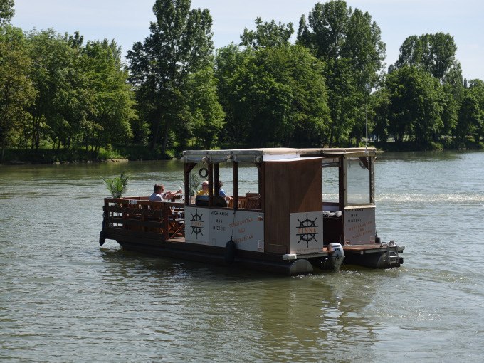 Ein rustikales Floßboot mit überdachtem Bereich fährt auf einem Fluss. Im Hintergrund sind grüne Bäume zu sehen., © Cool-Tours StattReisen
