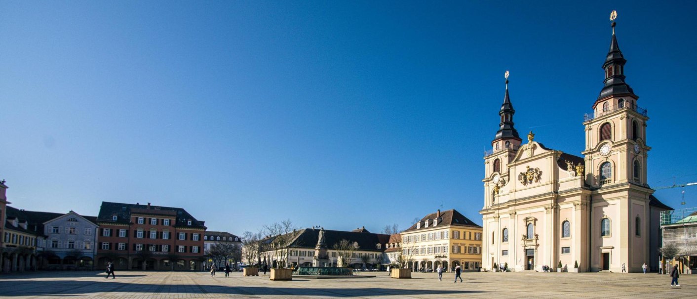 Der Ludwigsburger Marktplatz zeigt eine barocke Kirche und umliegende Gebäude unter klarem Himmel. Wenige Menschen sind auf dem Platz zu sehen., © SMG, Sarah Schmid Der Ludwigsburger Marktplatz zeigt eine barocke Kirche und umliegende Gebäude unter klarem Himmel. Wenige Menschen sind auf dem Platz zu sehen., © SMG, Sarah Schmid