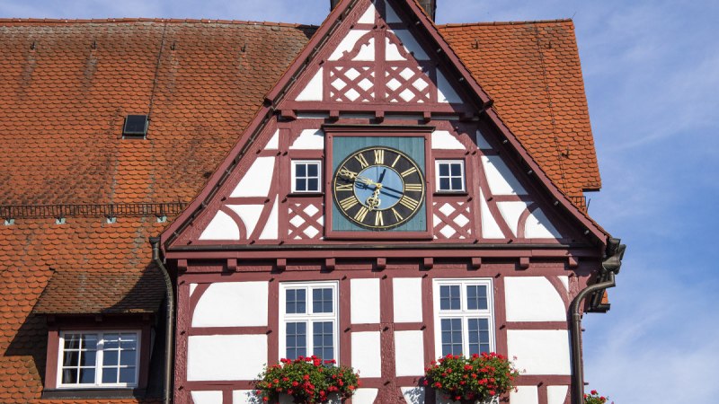 Fachwerk-Rathaus von Bad Urach mit Uhr und Blumenkästen, unter blauem Himmel. Das Gebäude hat ein rotes Dach und eine Turmspitze., © Bad Urach Tourismus