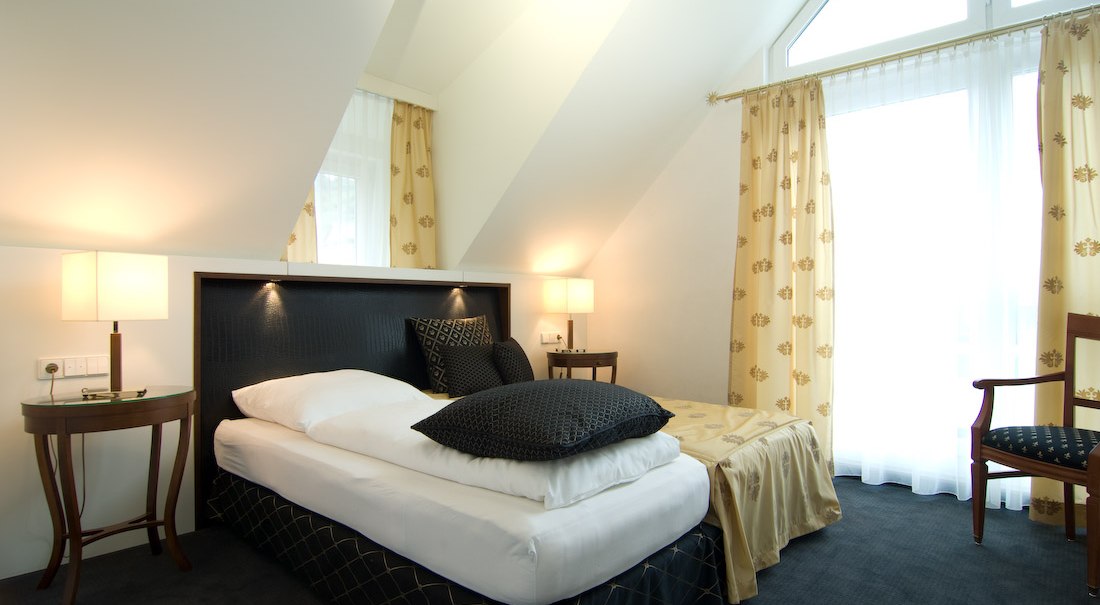 Elegantes Hotelzimmer mit Einzelbett, Nachttischen und großen Fenstern mit Vorhängen. Helle Wände und dunkle Akzente schaffen eine gemütliche Atmosphäre., © Jeanette Bender Elegantes Hotelzimmer mit Einzelbett, Nachttischen und großen Fenstern mit Vorhängen. Helle Wände und dunkle Akzente schaffen eine gemütliche Atmosphäre., © Jeanette Bender