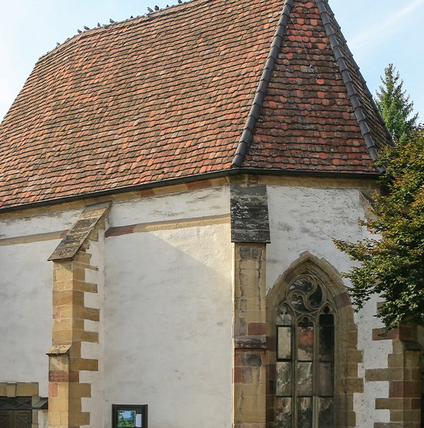 Historische Kirche mit rotem Ziegeldach, gotischem Fenster und Tauben auf dem Dach. Ein Baum und ein Schild sind ebenfalls sichtbar., © Alex Becher