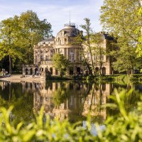 Schloss Monrepos spiegelt sich im ruhigen Wasser eines Sees, umgeben von &uuml;ppigem Gr&uuml;n und B&auml;umen. Menschen spazieren am Ufer entlang., &copy; SMG, Werner Dieterich