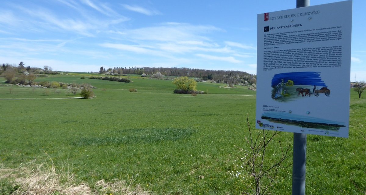 Ein Schild am Rutesheimer Grenzweg informiert über den Kastenbrunnen. Im Hintergrund erstreckt sich eine grüne Landschaft unter einem blauen Himmel., © Natur.Nah. Schönbuch & Heckengäu Ein Schild am Rutesheimer Grenzweg informiert über den Kastenbrunnen. Im Hintergrund erstreckt sich eine grüne Landschaft unter einem blauen Himmel., © Natur.Nah. Schönbuch & Heckengäu