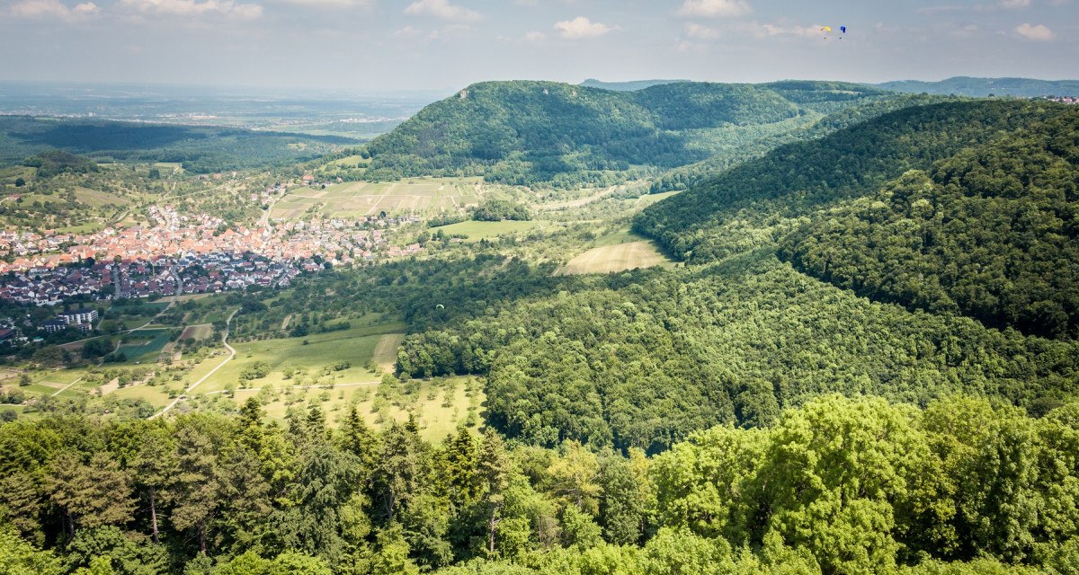 Blick von der Burg Hohenneuffen auf das Dorf Beuren, umgeben von grünen Wäldern und Hügeln unter einem blauen Himmel mit wenigen Wolken., © hochgehberge