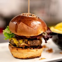 Ein saftiger Burger mit Salat und Käse auf einem Teller, daneben eine Schale mit Pommes frites., © Sensapolis GmbH