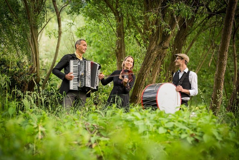 Drei Musiker mit Akkordeon, Geige und Trommel spielen in einem grünen Waldstück. Sie wirken fröhlich und entspannt., © Kulturhaus Schwanen