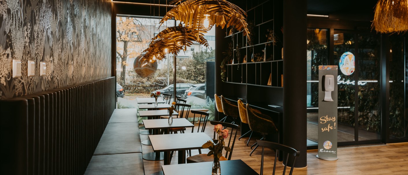 Moderne Lobby mit stilvollen Tischen und Stühlen, dekorativen Lampen und einem Desinfektionsmittelspender. Große Fenster bieten Blick nach draußen., © Rioca Stuttgart Posto 4 GmbH