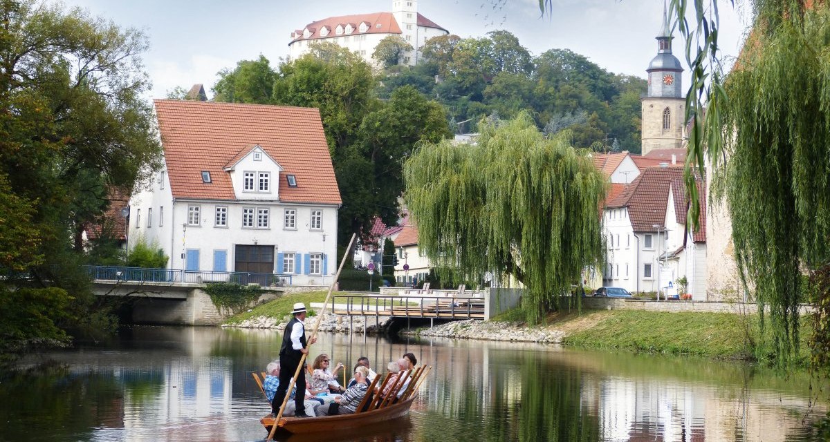 Ein Stocherkahn mit Passagieren fährt auf einem Fluss. Im Hintergrund sind eine Kirche, ein Schloss und malerische Häuser zu sehen. Ein Stocherkahn mit Passagieren fährt auf einem Fluss. Im Hintergrund sind eine Kirche, ein Schloss und malerische Häuser zu sehen.