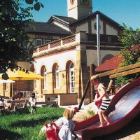 Kinder spielen auf einer Rutsche vor einem historischen Gebäude mit Turm und Sonnenschirmen im Garten., © Stuttgart-Marketing GmbH