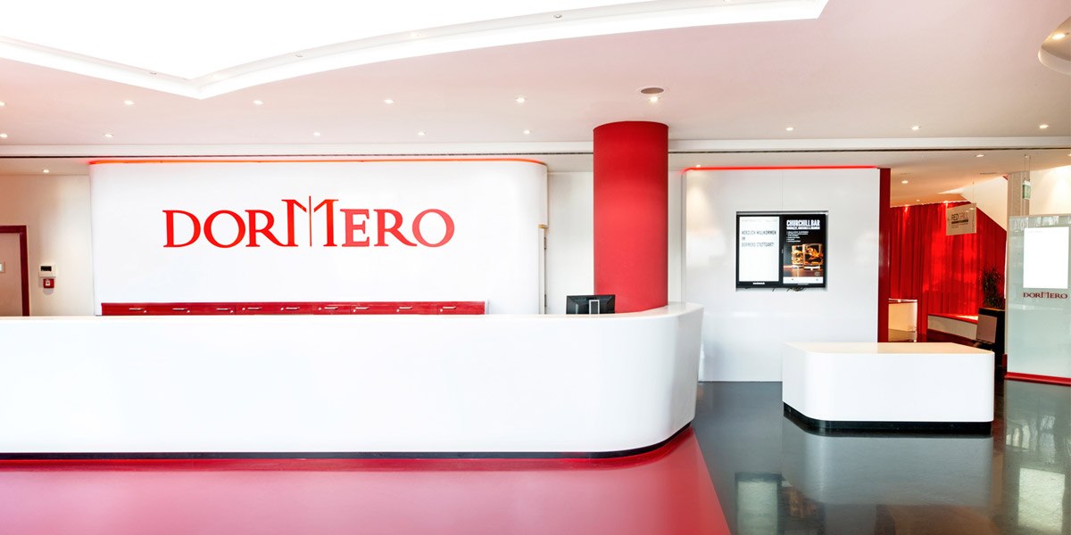 Moderne Hotelrezeption mit weiß-rotem Design, DORMERO-Logo an der Wand, Bildschirm mit Informationen, roter Teppichboden und helle Beleuchtung., © Dormero Hotel Stuttgart Moderne Hotelrezeption mit weiß-rotem Design, DORMERO-Logo an der Wand, Bildschirm mit Informationen, roter Teppichboden und helle Beleuchtung., © Dormero Hotel Stuttgart