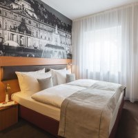 Modernes Hotelzimmer mit Doppelbett, großem Wandbild eines Gebäudes, Fenster mit weißem Vorhang und Wand-TV., © Novum Hospitality Modernes Hotelzimmer mit Doppelbett, großem Wandbild eines Gebäudes, Fenster mit weißem Vorhang und Wand-TV., © Novum Hospitality