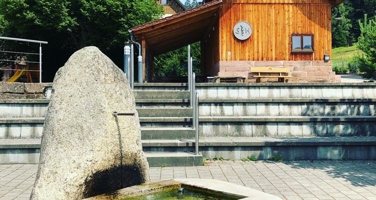 Steinbrunnen auf gepflastertem Platz, dahinter ein Holzgebäude mit Treppen und Bäumen. Klare, sonnige Atmosphäre., © Nördlicher Schwarzwald Steinbrunnen auf gepflastertem Platz, dahinter ein Holzgebäude mit Treppen und Bäumen. Klare, sonnige Atmosphäre., © Nördlicher Schwarzwald