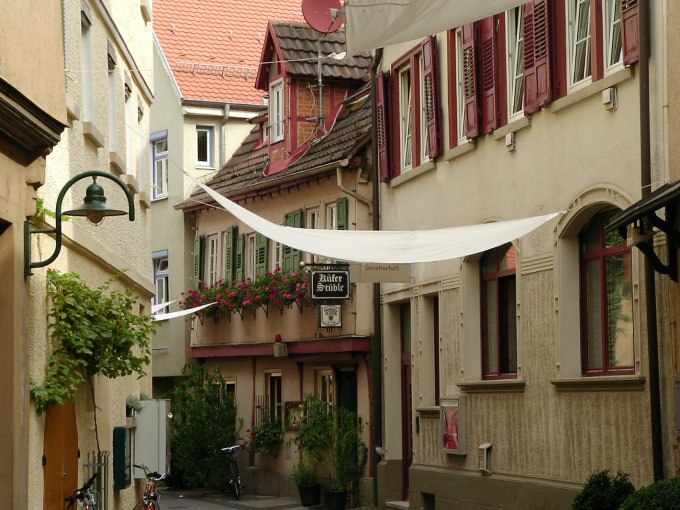 Schmale Gasse mit historischen Gebäuden, roten Fensterläden und Blumen. Ein Schild weist auf ein Restaurant hin. Weiße Tücher spannen sich über die Straße., © Cool-Tours StattReisen Schmale Gasse mit historischen Gebäuden, roten Fensterläden und Blumen. Ein Schild weist auf ein Restaurant hin. Weiße Tücher spannen sich über die Straße., © Cool-Tours StattReisen