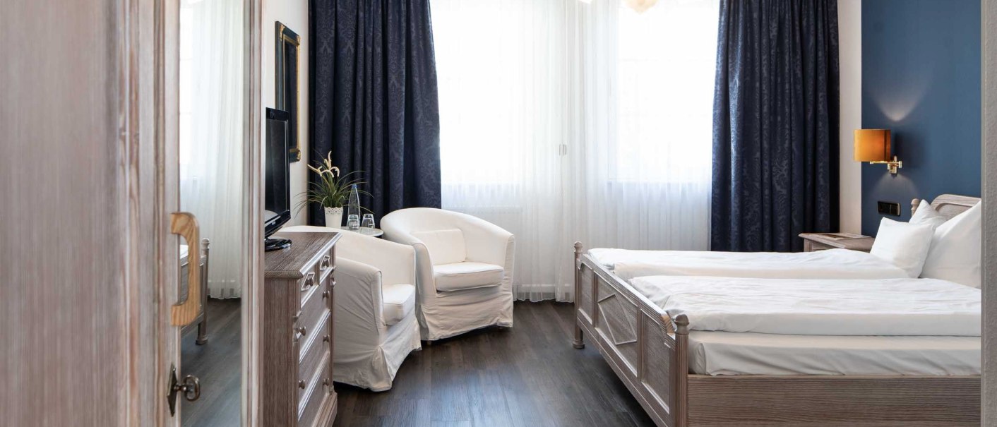 Elegantes Hotelzimmer mit Doppelbett, weißen Sesseln, dunklen Vorhängen und Holzmöbeln. Helle Beleuchtung und stilvolle Dekoration., © Flairhotel Vier Jahreszeiten