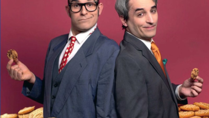 Zwei M&auml;nner in Anz&uuml;gen stehen R&uuml;cken an R&uuml;cken vor einem Haufen Kekse. Sie halten jeweils einen Keks in der Hand und l&auml;cheln verschmitzt., &copy; Theaterwerkstatt Schw&auml;bisch Gm&uuml;nd e.V.