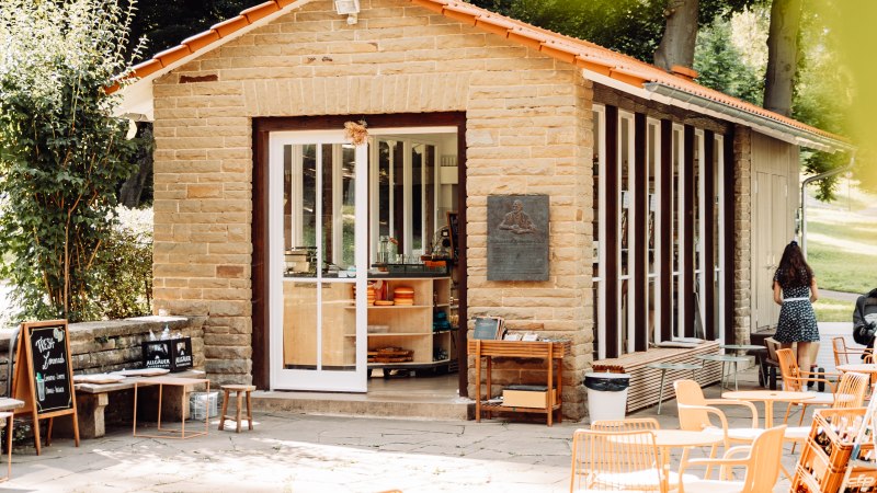 Kleines Caf&eacute; mit Steinw&auml;nden und gro&szlig;en Fenstern, umgeben von B&auml;umen. Orangefarbene St&uuml;hle und Tische auf der Terrasse, einladende Atmosph&auml;re., &copy; L. Prestle/ Documenting Love