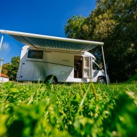 Ein Wohnmobil mit ausgefahrener Markise steht auf einer gr&uuml;nen Wiese unter blauem Himmel. Im Hintergrund sind B&auml;ume und ein Geb&auml;ude zu sehen., &copy; SMG, Thomas Niederm&uuml;ller