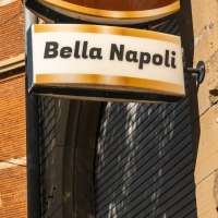 Ein Schild mit der Aufschrift 'Bella Napoli' hängt an einem Backsteingebäude. Der Himmel ist klar und blau, ein Baum ist im Vordergrund sichtbar., © SMG, Sarah Schmid