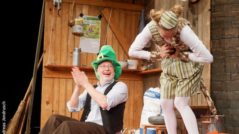 Ein Schauspieler mit gr&uuml;nem Hut lacht und klatscht, w&auml;hrend eine andere Person in gestreiftem Kost&uuml;m sich verbeugt. Holzwand im Hintergrund., &copy; Stadt Kornwestheim Kulturmanagement