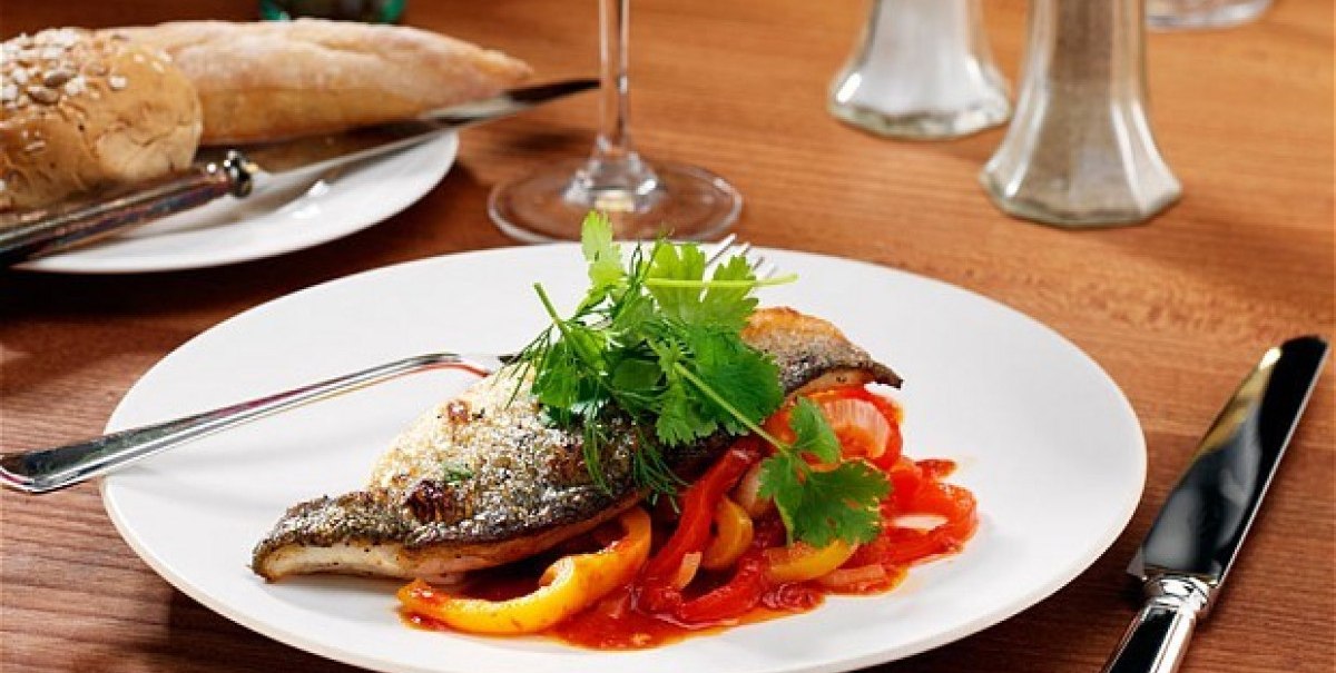 Fischfilet auf Paprika mit frischen Kräutern, serviert auf einem weißen Teller. Im Hintergrund ein Glas Wein und Brot auf einem Holztisch.