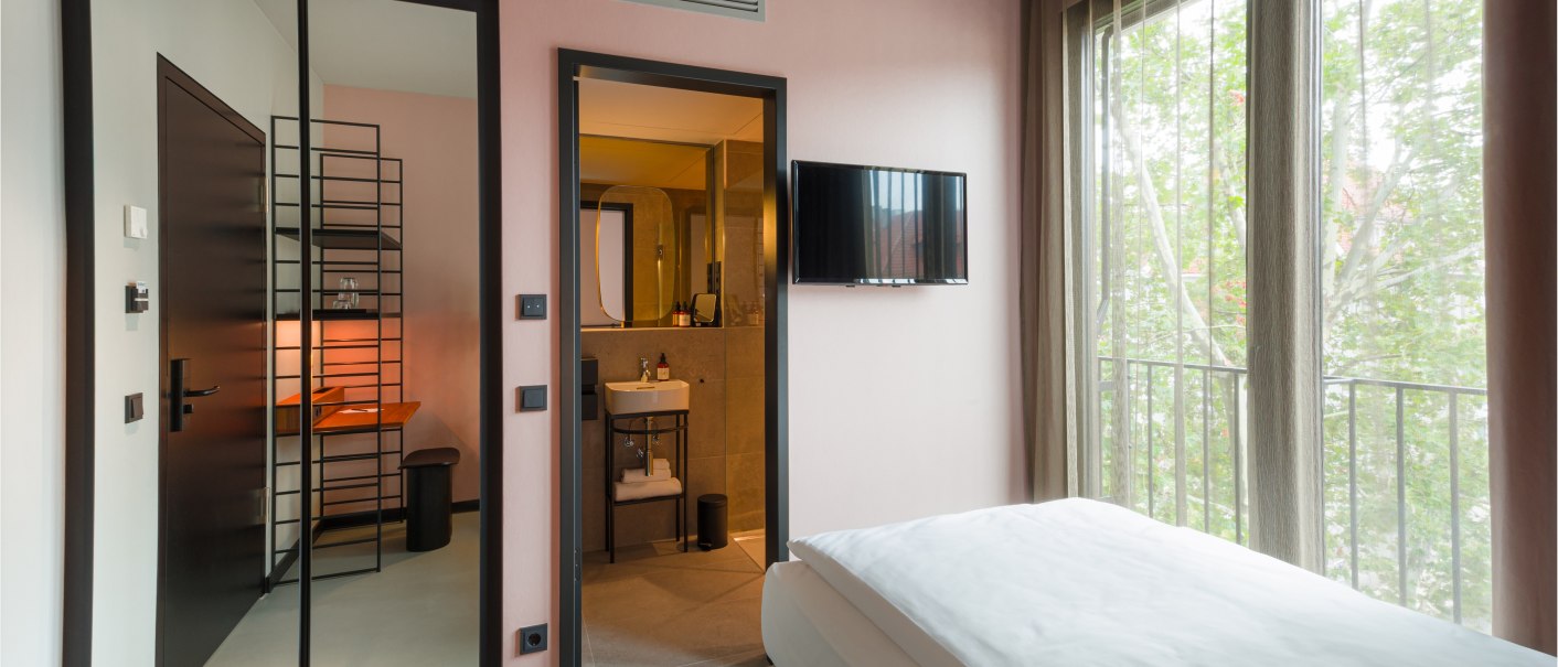 Modernes Zimmer mit Bett, Fernseher an der Wand, großem Fenster mit Vorhängen, Spiegel und offenem Badezimmer mit Waschbecken., © EmiLu Hotel GmbH Modernes Zimmer mit Bett, Fernseher an der Wand, großem Fenster mit Vorhängen, Spiegel und offenem Badezimmer mit Waschbecken., © EmiLu Hotel GmbH