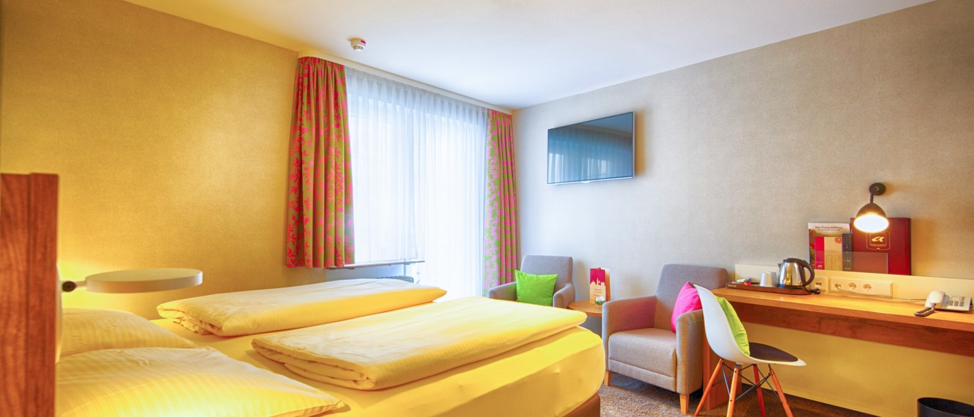Ein gem&uuml;tliches Hotelzimmer mit Doppelbett, Schreibtisch, Fernseher und zwei Sesseln. Helle Vorh&auml;nge und bunte Kissen sorgen f&uuml;r eine einladende Atmosph&auml;re., &copy; TOMAS