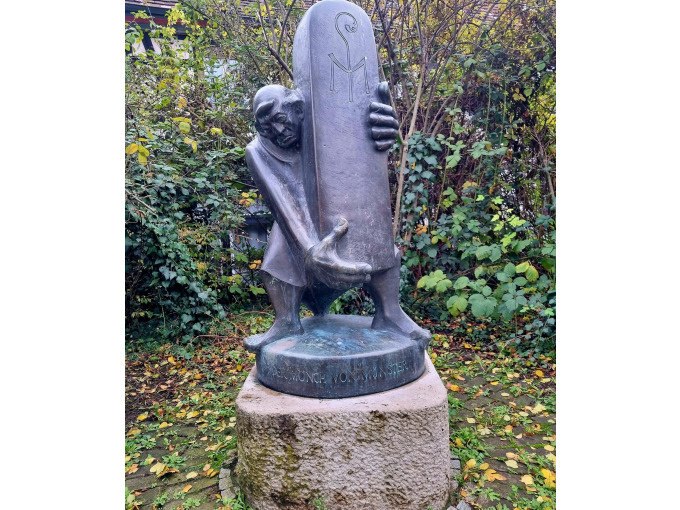 Bronzestatue eines Mannes, der eine gro&szlig;e Steintafel umarmt, auf einem Sockel im Freien, umgeben von Laub und B&uuml;schen., &copy; Cool-Tours StattReisen