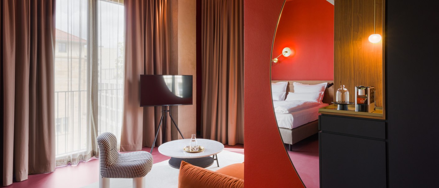 Moderne Hotelsuite mit roten Wänden, gemütlichem Bett, Sitzbereich mit Sessel und Tisch, Fernseher und stilvoller Beleuchtung., © Emilu Hotel GmbH Moderne Hotelsuite mit roten Wänden, gemütlichem Bett, Sitzbereich mit Sessel und Tisch, Fernseher und stilvoller Beleuchtung., © Emilu Hotel GmbH