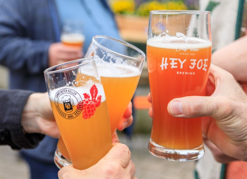 Drei Personen stoßen mit Biergläsern an, die Logos von "Hey Joe Brewing" und "Cannstatter Keller Bräu" tragen.