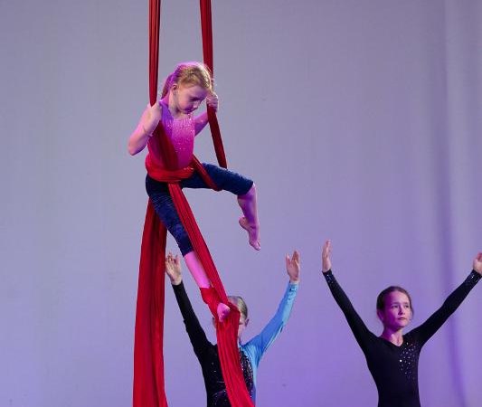 Ein Kind zeigt Luftakrobatik an roten Tüchern, während zwei andere Kinder auf dem Boden knien und die Arme heben., © Kunstschule Labyrinth Ein Kind zeigt Luftakrobatik an roten Tüchern, während zwei andere Kinder auf dem Boden knien und die Arme heben., © Kunstschule Labyrinth