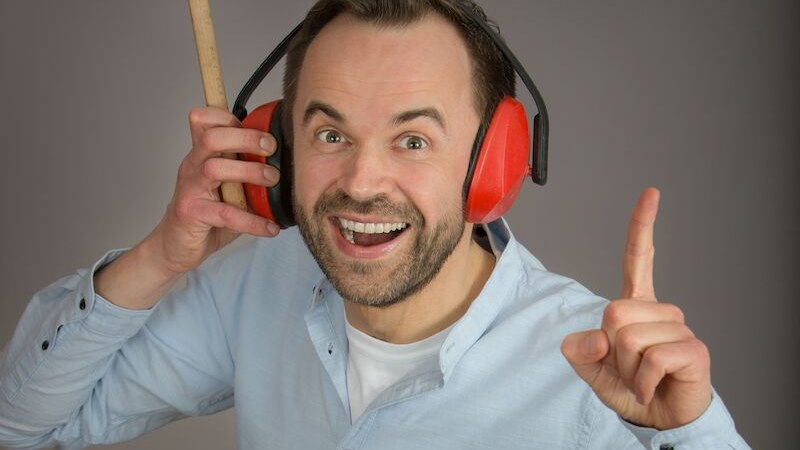 Ein Mann mit roten Ohrensch&uuml;tzern h&auml;lt einen Hammer ans Ohr und l&auml;chelt, w&auml;hrend er mit dem Finger nach oben zeigt. Er tr&auml;gt ein hellblaues Hemd., &copy; Kurverwaltung Bad Urach