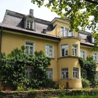 Eine gelbe Villa mit Efeu bewachsen, umgeben von einem gepflegten Garten. Die Architektur ist klassisch mit mehreren Fenstern und einem kleinen Balkon., &copy; cc_Ruoff Stiftung