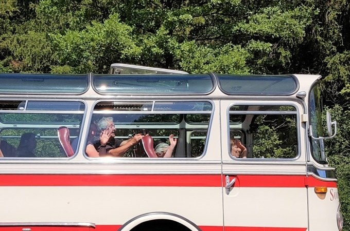 Ein Oldtimerbus mit roten und weißen Streifen fährt durch eine grüne Landschaft. Die Passagiere winken fröhlich aus den Fenstern., © SwabianTravel