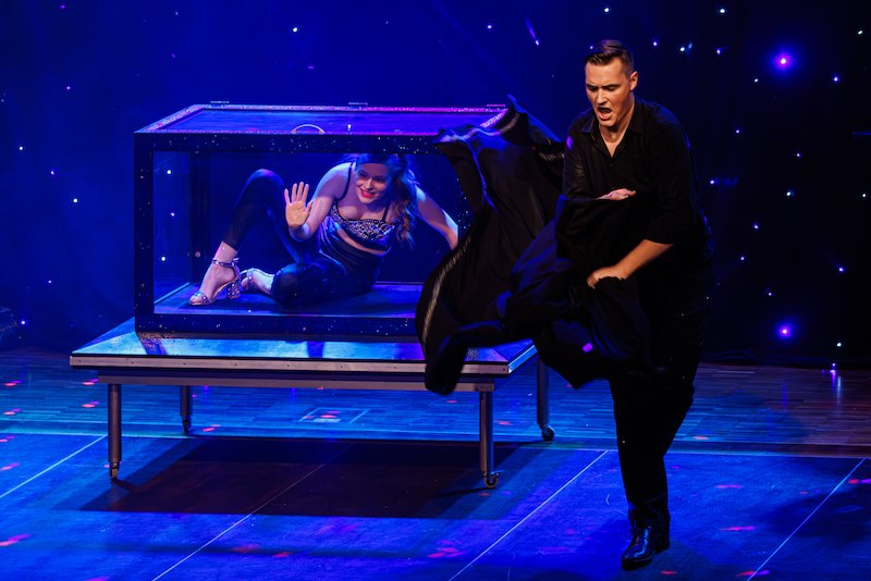 Eine Frau sitzt in einem Glaskasten auf der Bühne, während ein Mann mit einem schwarzen Umhang eine Zaubershow vorführt., © Scala Kultur Live gGmbH Eine Frau sitzt in einem Glaskasten auf der Bühne, während ein Mann mit einem schwarzen Umhang eine Zaubershow vorführt., © Scala Kultur Live gGmbH