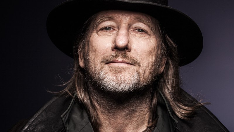 Mann mit langem Haar, schwarzem Hut und Lederjacke blickt in die Kamera vor einem dunklen Hintergrund., © Mauerwerk GmbH Mann mit langem Haar, schwarzem Hut und Lederjacke blickt in die Kamera vor einem dunklen Hintergrund., © Mauerwerk GmbH