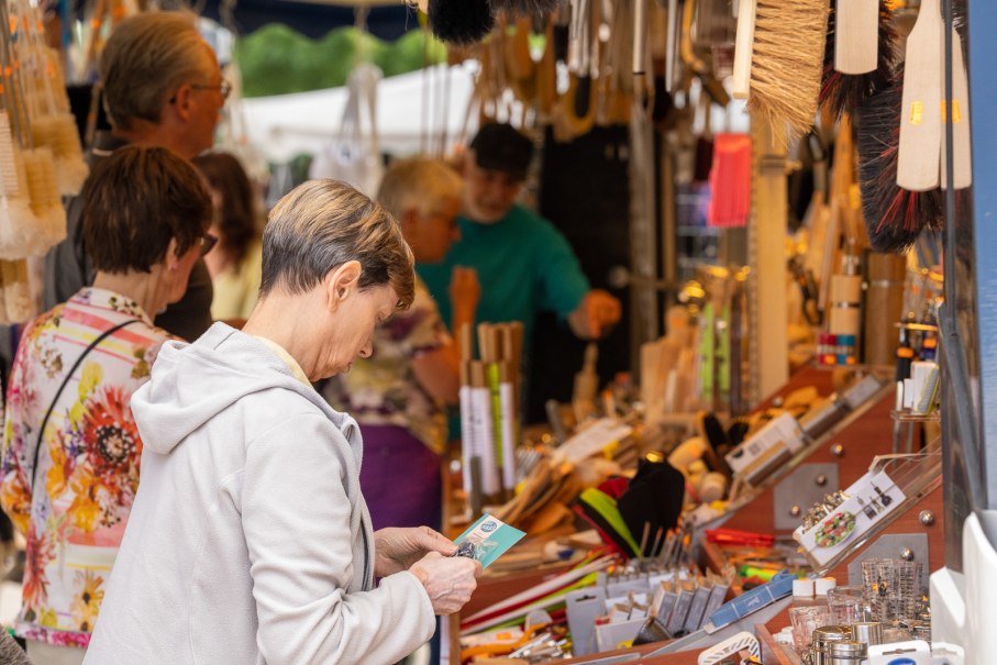 Menschen stöbern an einem Marktstand voller Bürsten und Haushaltswaren auf einem Sommermarkt in Nürtingen., © Jürgen Hackenjos Menschen stöbern an einem Marktstand voller Bürsten und Haushaltswaren auf einem Sommermarkt in Nürtingen., © Jürgen Hackenjos