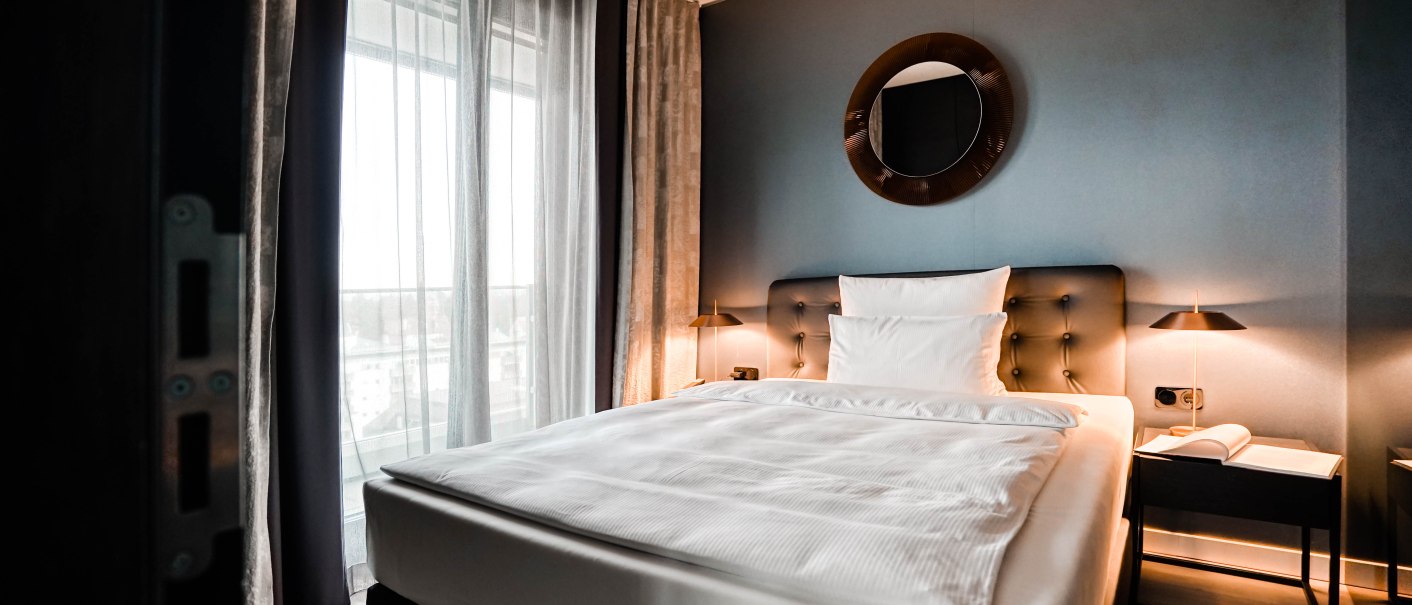 Modernes Hotelzimmer mit großem Bett, runden Spiegel an der Wand, Nachttischlampen und bodenlangen Vorhängen vor einem Fenster., © CLOUD N°7 APARTMENTS GmbH