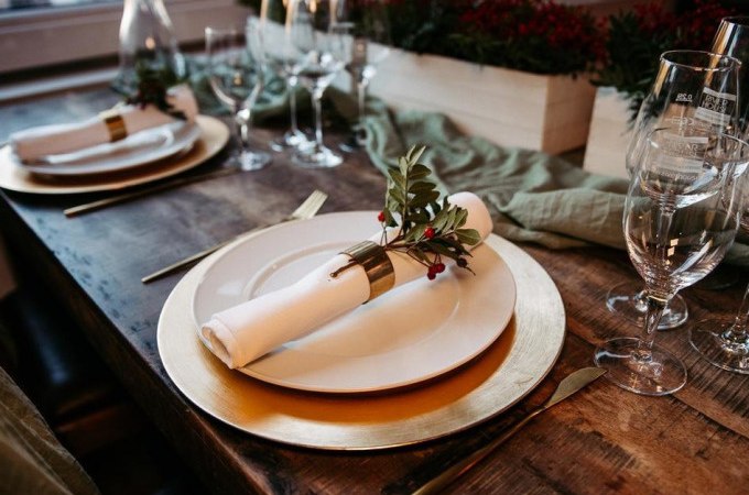 Elegante Tischdekoration mit goldenen Tellern, weißen Servietten und grünen Zweigen auf einem Holztisch, umgeben von Weingläsern., © Cool-Tours StattReisen