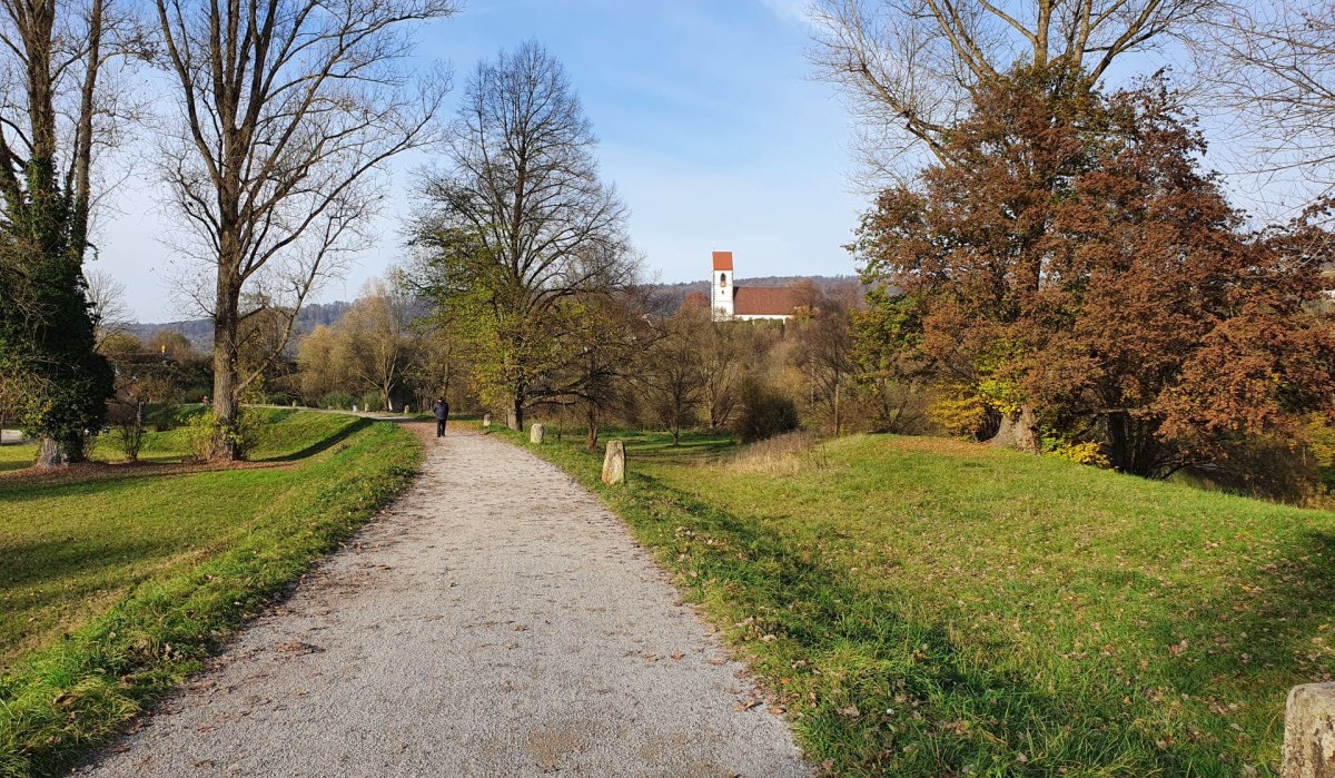 Ein Kiesweg führt durch einen grünen Park mit kahlen Bäumen. Im Hintergrund ist eine Kirche mit rotem Dach zu sehen., © Kulturamt Plochingen Ein Kiesweg führt durch einen grünen Park mit kahlen Bäumen. Im Hintergrund ist eine Kirche mit rotem Dach zu sehen., © Kulturamt Plochingen