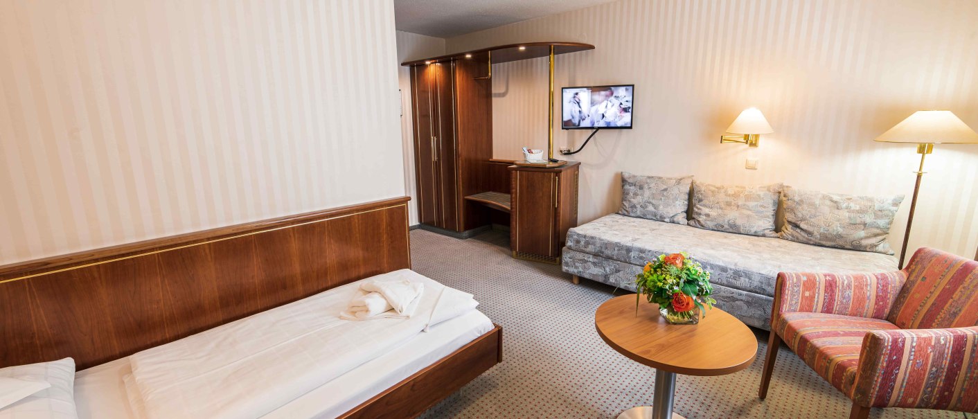Ein gemütliches Hotelzimmer mit Einzelbett, Sofa, Sessel, Tisch mit Blumen und einem Fernseher an der Wand., © TOMAS