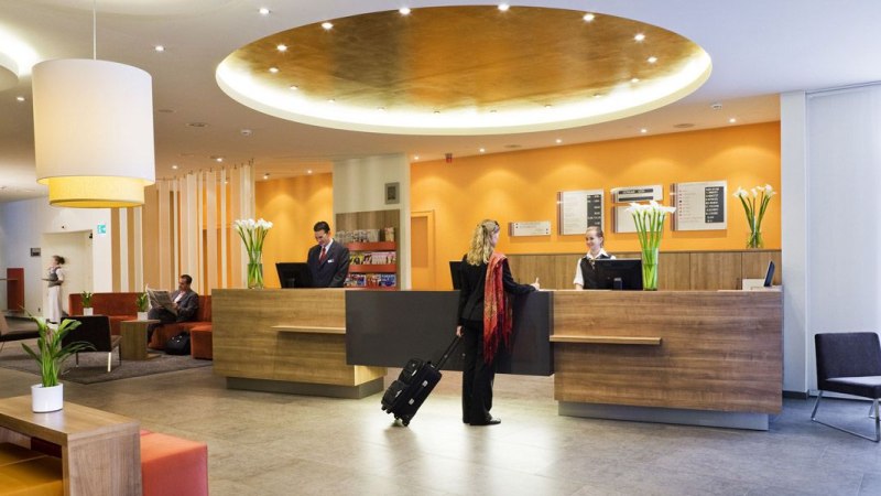 Eine moderne Hotelrezeption mit zwei Angestellten und einer Frau mit Koffer. Im Hintergrund sitzen G&auml;ste auf Sofas. Die Einrichtung ist stilvoll und einladend., &copy; Mercure Stuttgart Airport Messe