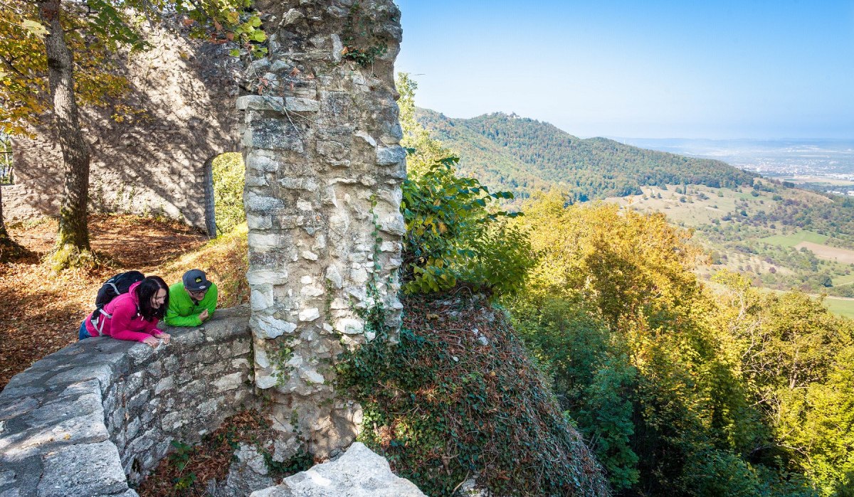 Zwei Personen in bunter Kleidung betrachten eine alte Steinmauer in einer bewaldeten Landschaft mit weitem Ausblick., © hochgehberge