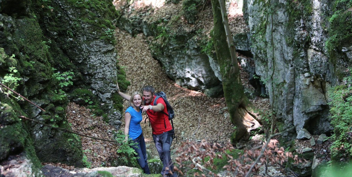 Zwei Wanderer in bunter Kleidung erkunden eine grüne, felsige Schlucht mit Moos und Laub bedeckt.