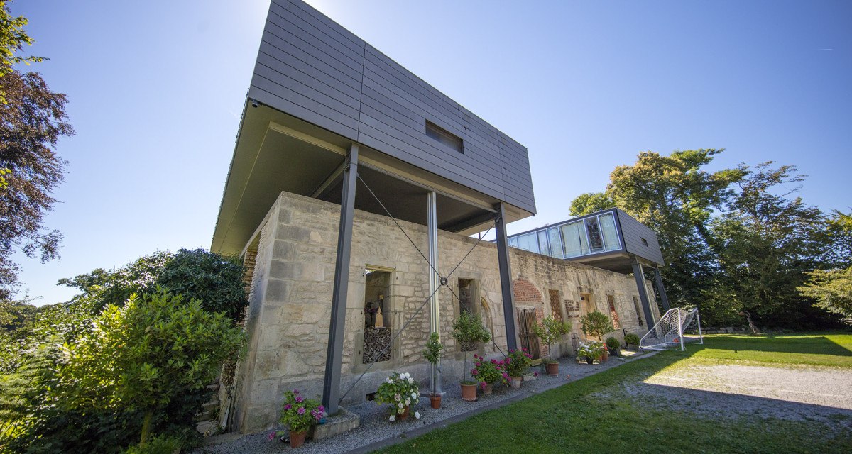 Modernes Gebäude mit alten Steinmauern und Glasstruktur, umgeben von Pflanzen und Bäumen unter blauem Himmel., © Region Stuttgart